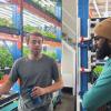 Two people stand in front of plants that are growing beneath lights while indoors.