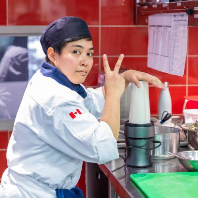 A person wearing a chef outfit flashes the peace sign as they use a blender in a kitchen.