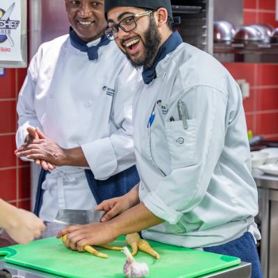 Two smiling people wearing chef outfits prepare vegetables on a cutting board at a kitchen station.