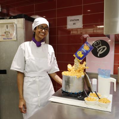 A person wearing a chef's uniform stands next to a cake shaped to look like Kraft Dinner.
