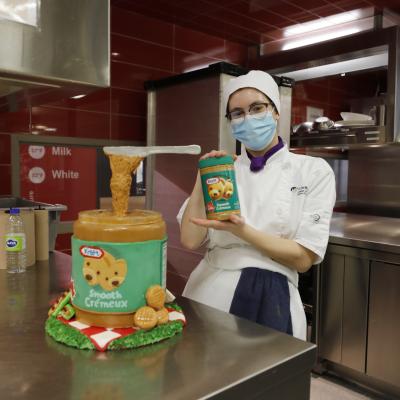 A person wearing a chef's uniform stands next to a cake shaped to look like a jar or peanut butter.