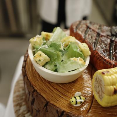 A cake made to look like a barbecued steak with Caesar salad and corn on the cob.