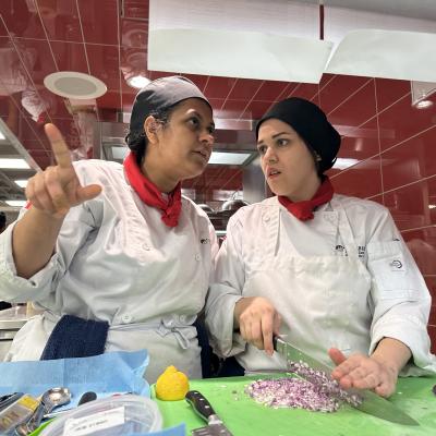 Two people wearing chef’s uniforms dice ingredients in a kitchen.