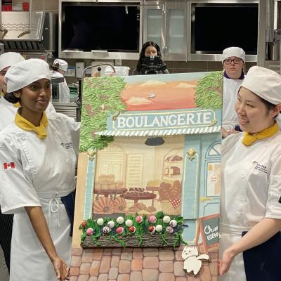 Two people wearing chef outfits hold up a painting of a French bakery that has edible elements to it.