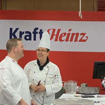 Two people wearing chef’s uniforms chat while standing in front of a Kraft Heinz sign.
