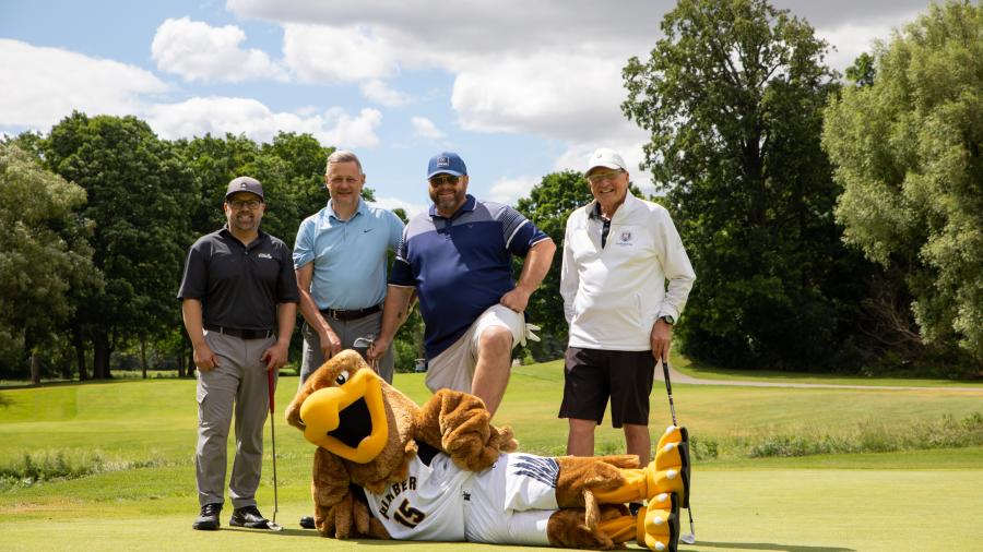 Four men stand in a row wearing golf clothes and holding golf clubs. The Humber Hawk mascot lies on the ground in front of them.