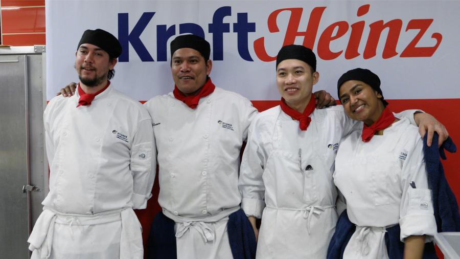 Four individuals stand arm-in-arm wearing chef uniforms.