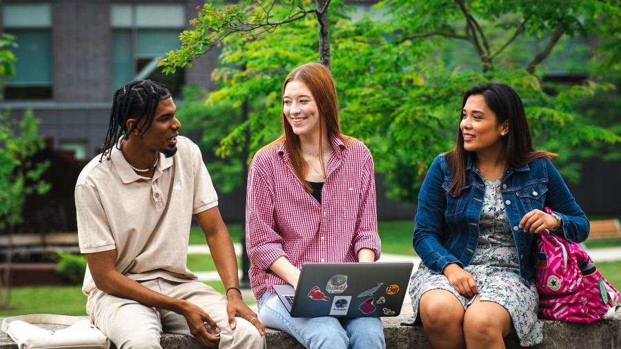 Three people sit together outdoors. One has a laptop resting on their lap.