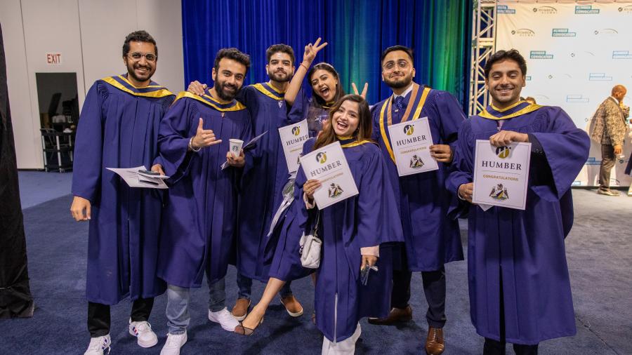 A group of students wearing graduation gowns smile and pose for a photo while holding up signs that say Humber and congratulatio