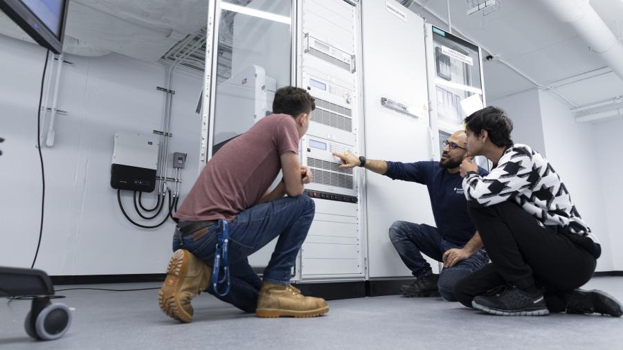 A person points at an electrical device while two other people crouch down to look.