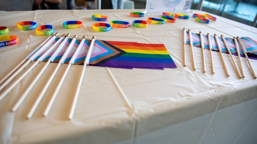 Several Pride and Trans flags rest on a table that has rainbow-coloured bracelets on it.