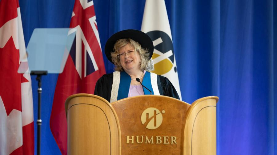 A person wearing a gap and graduation gown speaks while standing at a podium with the Humber College logo on it