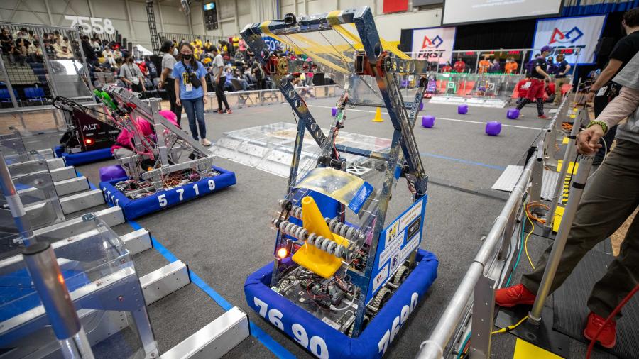 Several robots are lined up inside an arena while spectators sit in the stands.