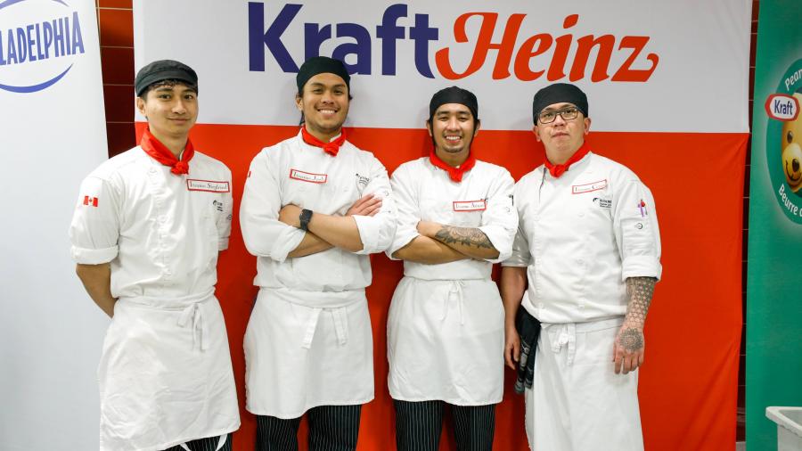 Four smiling people wearing chef’s outfits stand in front of a sign that reads Kraft Heinz.