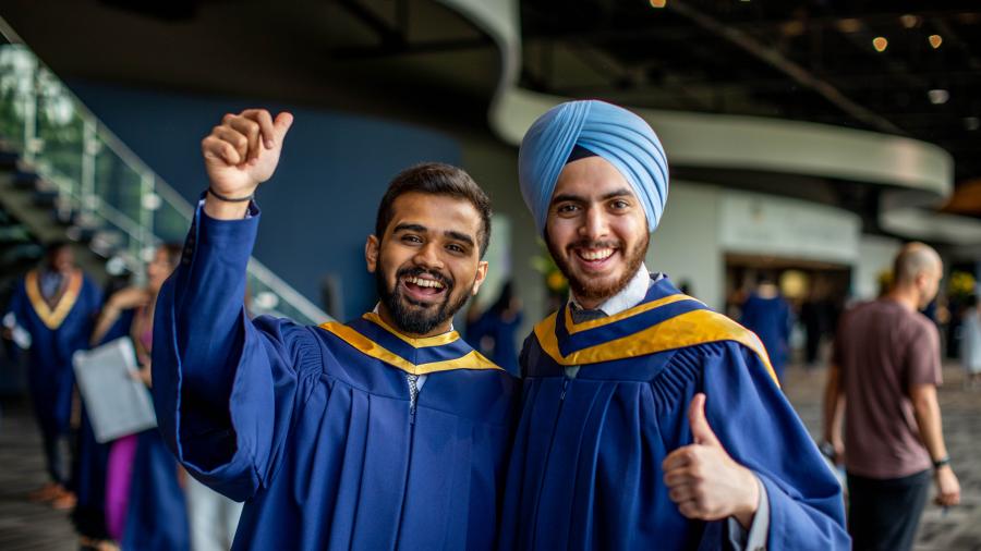 Two people wearing graduation robes smile and give the thumb’s up as they pose for a photo.