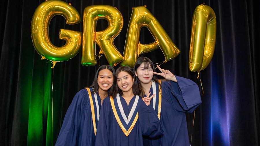 Three people wearing graduation gowns smile and pose for a photo beneath a sign that reads ‘grad.’