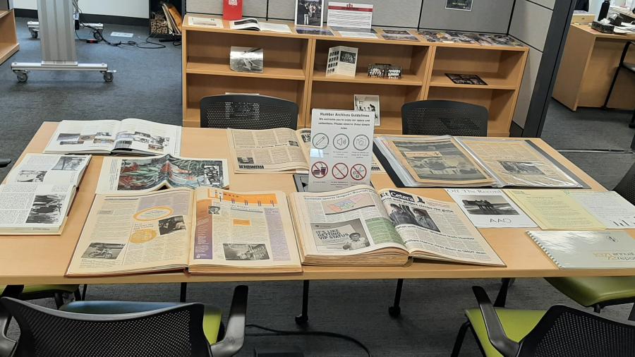 Bound copies of old newspapers sit on a desk. Photographs can be seen in the background on another table.