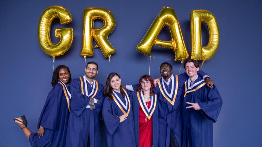 Several people wearing graduation gowns stand beneath a series of ballons spelling out the word Grad.