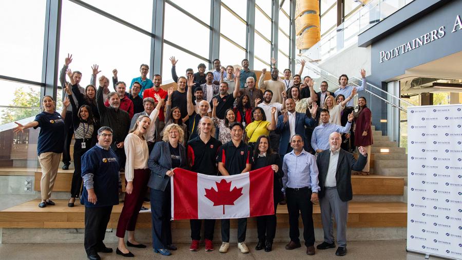 A large group of smiling and cheering people stand together. Two people at the front hold a Canadian flag.