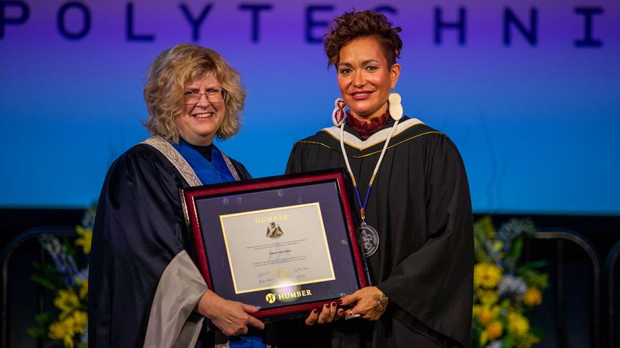 Two smiling people stand together wearing graduation gowns while holding a degree.