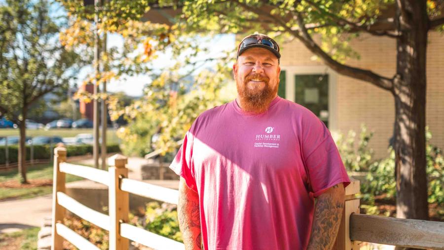 Scott Iles wears a pink shirt and poses in front of a fence and some foliage.