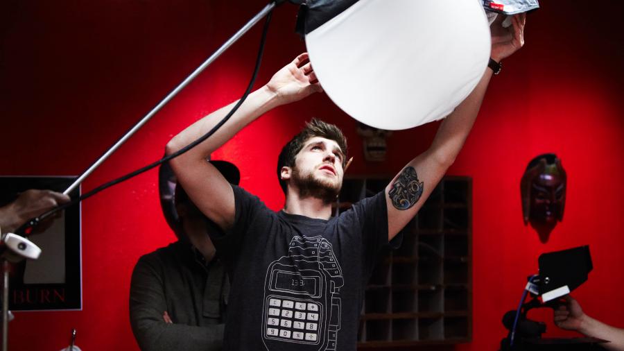 A young man holds a large, round, white set light over his head, shining into a red-walled studio.
