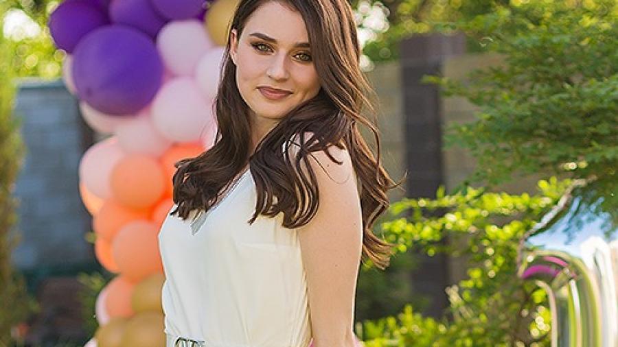 Renata Borsuk smiles softly at the camera, looking over her shoulder with balloons in the background wearing a white dress