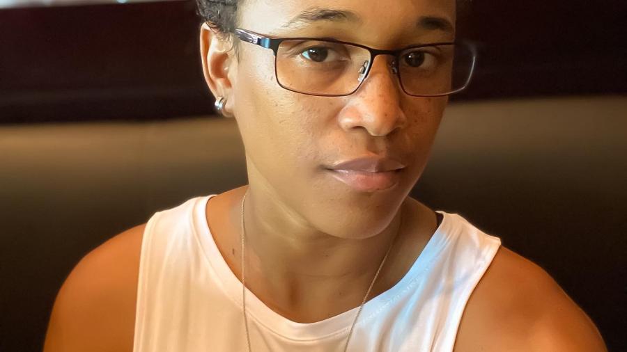 Krystal Moodie is seated, wearing a white sleeveless t-shirt and thin-framed glasses. She gazes sideways at the camera.