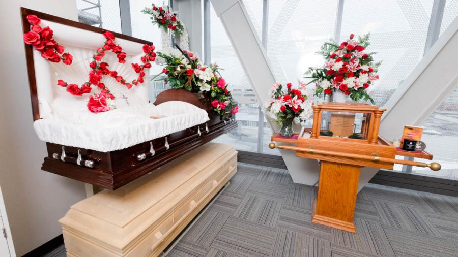 An open wood casket with soft white lining sits on a stand in front of a flower-covered altar in the funeral services lab