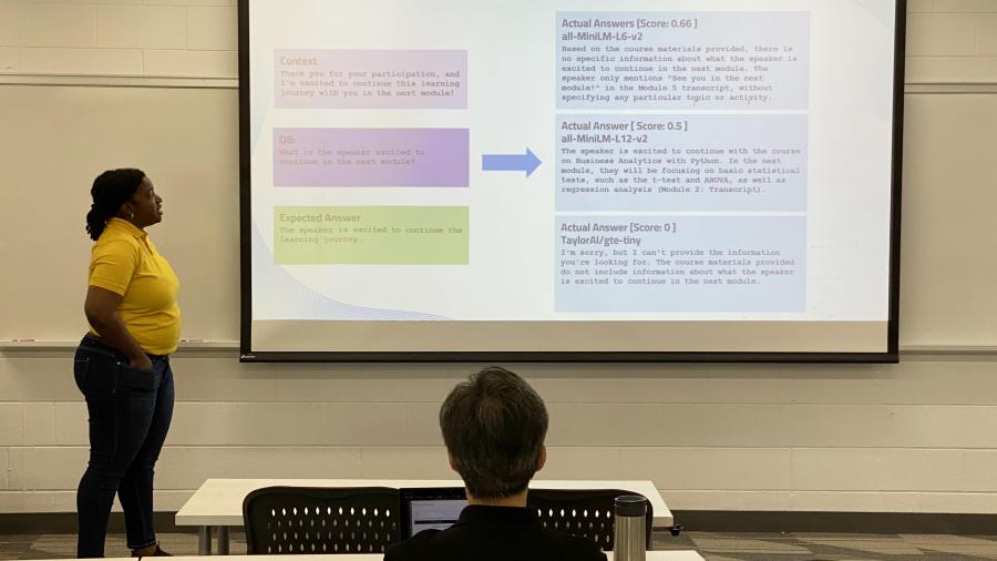 A person stands in front of a screen and gives a presentation while a professor listens.