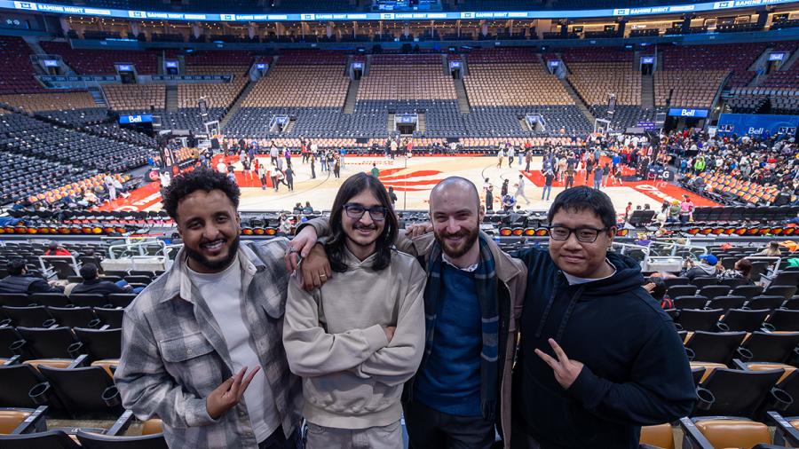 Four people stand together in a stadium. Behind them is the Toronto Raptors basketball court.