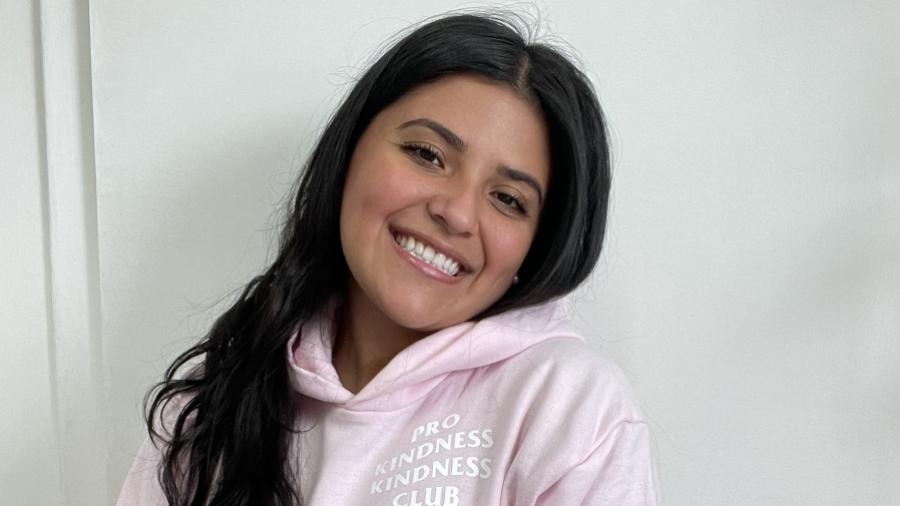 Camila Ruiz Tacha has long brown hair and one hand in the pocket of her pink sweatshirt and smiles, head tilted to the side.