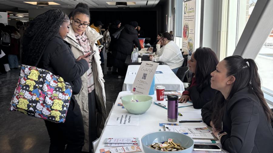 Two people stand in front of a table that has two other people sitting at it. The table has literature and other items on it.