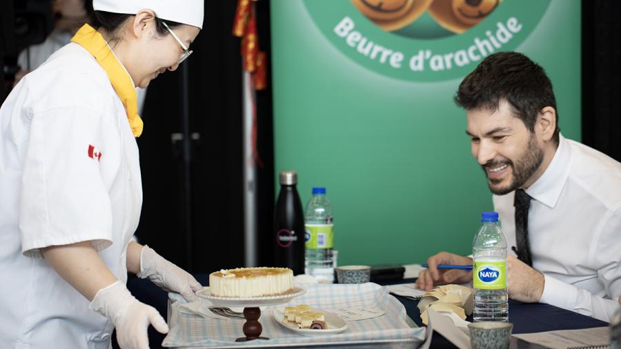 A person wearing a chef’s uniform presents plated dishes to a judge to sample.