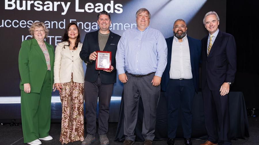 Several people stand together on a stage. One is holding an award.