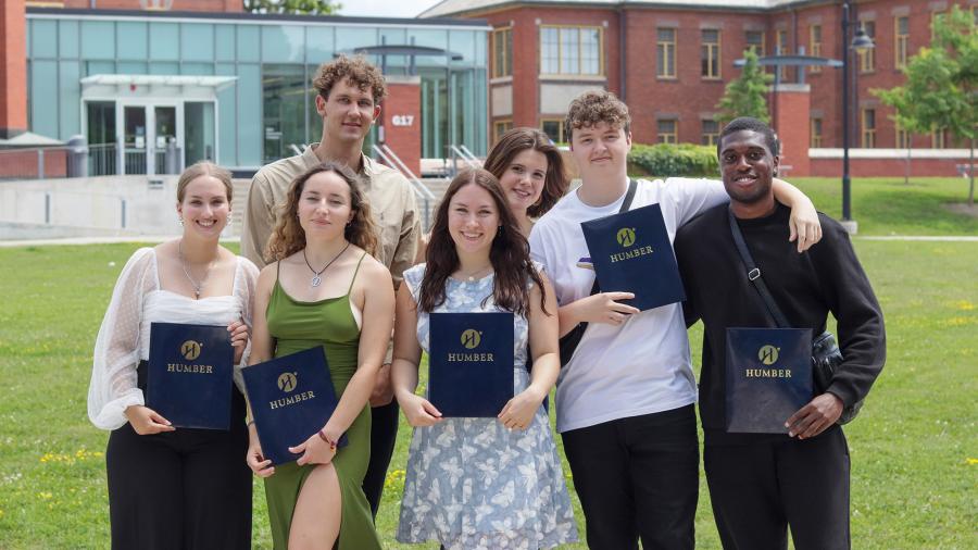 Seven people smile and pose for a photo while holding up blue folders with Humber written on them.
