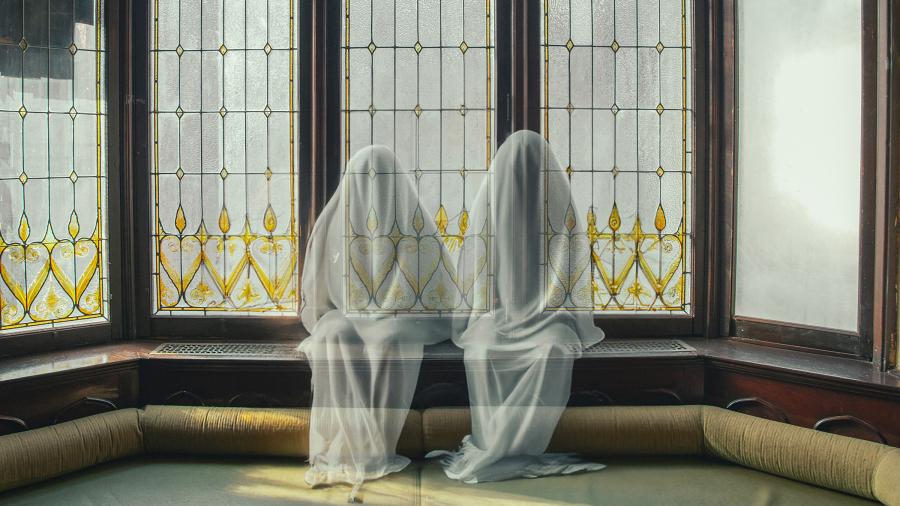 Two people dressed likes translucent ghosts sit in front of a stained glass window.