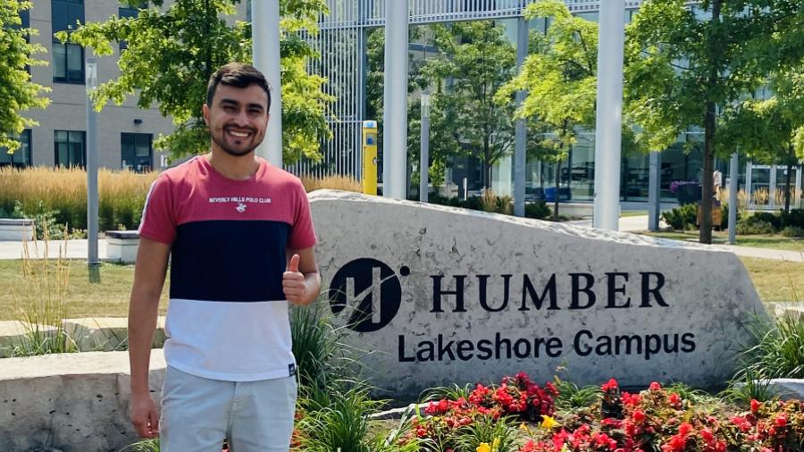 A smiling person stands in front of a sign the reads Humber Lakeshore Campus that sits in a flower bed.
