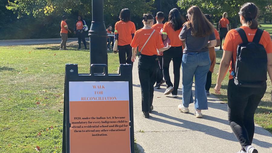People wearing orange shirts walk past a sign with Walk for Reconciliation on it along with other information.