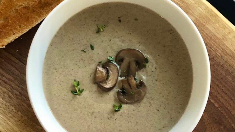 A bowl of mushroom soup sits on a cutting board beside bread and raw mushrooms. It is light brown, topped with brown mushrooms
