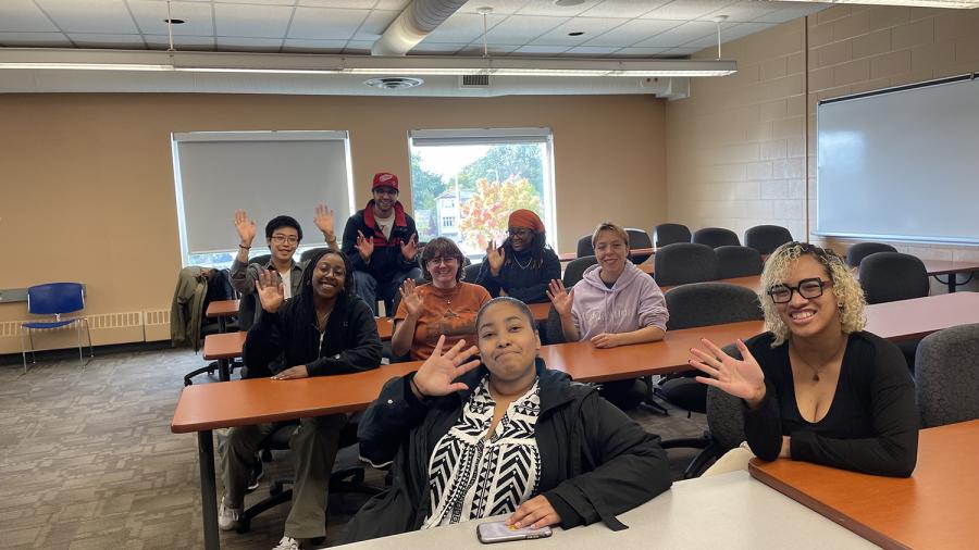 A group of people sit at desks while waving to the camera.