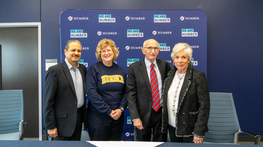 Four people smile and pose for a photo while standing in front of a blue banner with the Humber College logo on it. 
