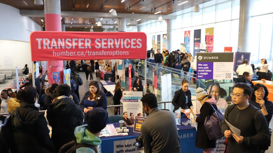 People walk through a room and stop to look at information being displayed on tables. A sign at the front reads Transfer Service