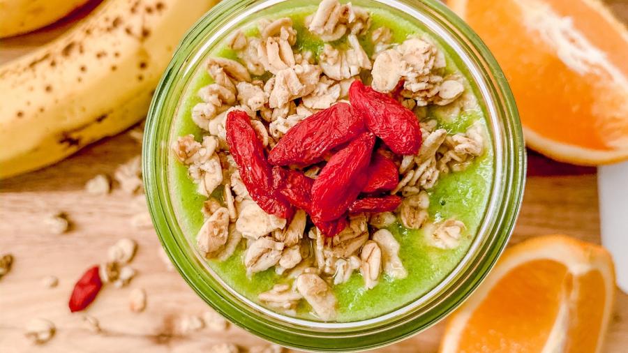 A green smoothie garnished with nuts and raspberries sits on a cutting board surrounded by orange slices and bananas