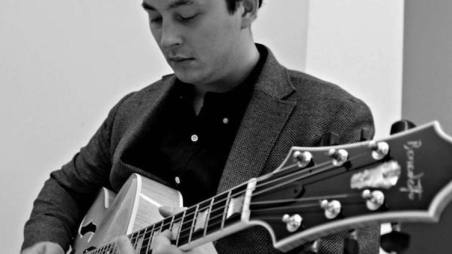 Lucian Gray is standing, wearing a blazer while playing guitar in a black and white photo.