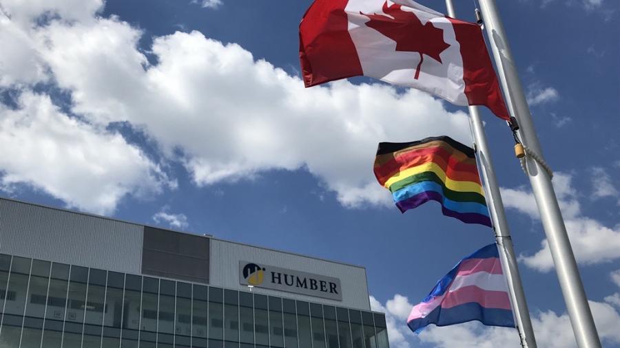 The Trans, Pride and Canadian flags are at half mast against a blue sky with puffy white clouds in front of Humber North's LRC
