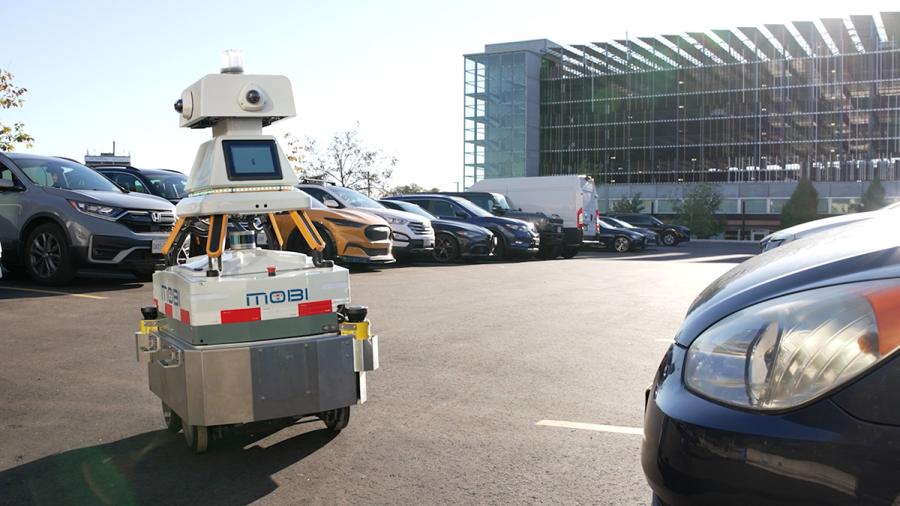 A wheeled robot is in a parking lot with cars.