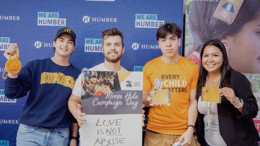 Four people stand together. One is holding up a sign that reads Moose Hide Campaign Day - Love is not Abuse.
