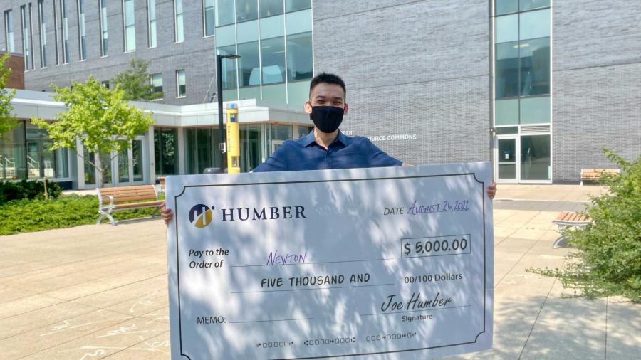 Newton Lew holds up a giant cheque for $5,000 from Humber College which hides most of his body.He's wearing a t-shirt and shorts