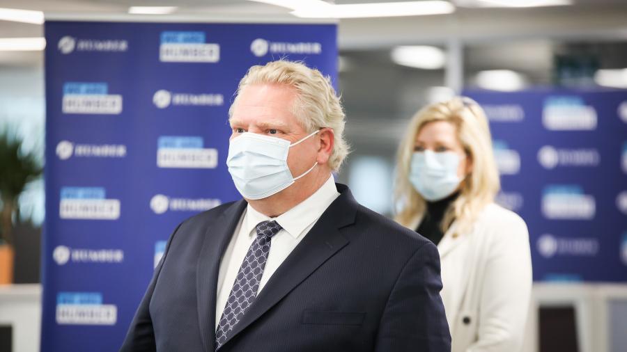 Premier Doug Ford wears a blue surgical mask at a press conference. MP Natalia Kusendova is seen in the background.
